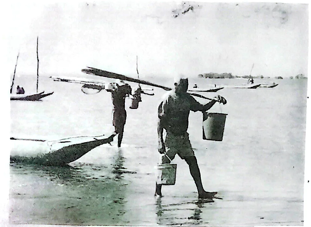 Pescadores caminhando nas águas rasas de Baiacu com baldes e embarcações ao fundo